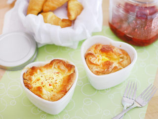 Two heart-shaped ham and swiss baked egg souffles are on a green placemat. A basket of croissants and jam sit behind them.