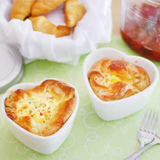 Two heart-shaped ham and swiss baked egg souffles are on a green placemat. A basket of croissants and jam sit behind them.