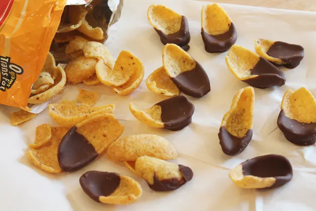 Frito corn chips pouring out of a bag on a sheet of parchment paper. Each chip has been half dipped in chocolate.