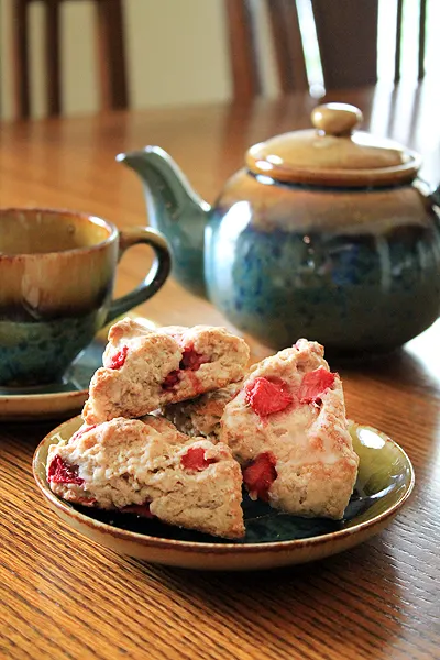 Three strawberry scones sit on a plate. A blue teapot and cup sit in the background.